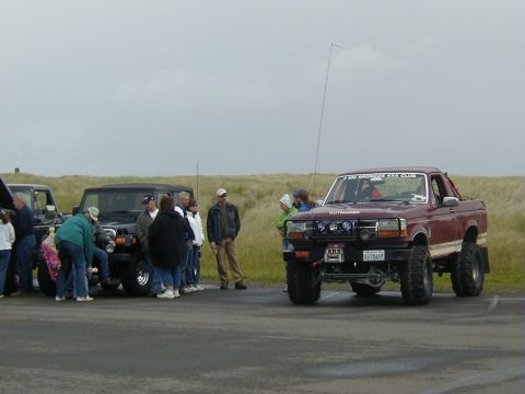 1994 Ford Explorer - A group of people stand around several off-road vehicles parked on a paved surface with grassy dunes in the background.