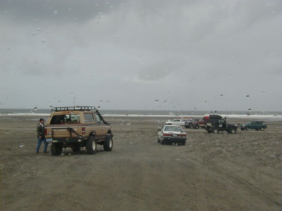 Tan lifted Ford 4x4 with roof rack parked on an overcast beach with other rigs