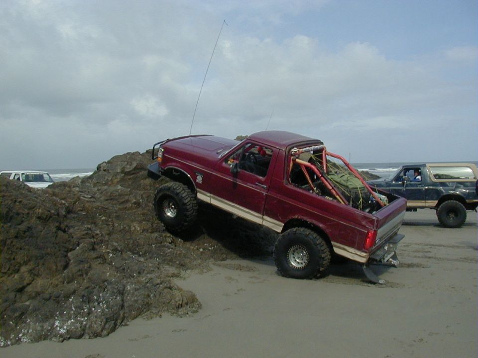 Lifted maroon Ford Bronco with roll cage climbing a rock outcrop on a beach