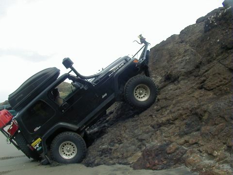 1994 Ford Explorer - A black Jeep Wrangler Rubicon navigates a steep, rocky terrain near a sandy beach, equipped for off-road adventure.