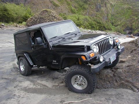 1994 Ford Explorer - A black Jeep Wrangler is parked on a sandy and rocky shore with vegetation on the hillside.
