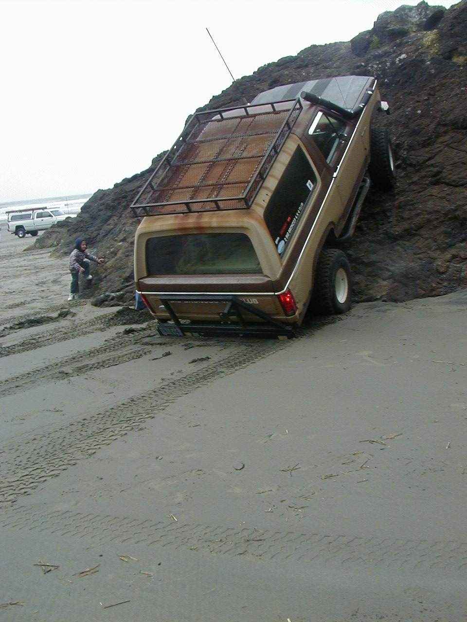 Brown SUV with roof rack climbing a rocky beach incline. Another SUV parked on the sand nearby