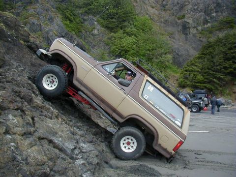 1994 Ford Explorer - A modified Ford Bronco navigates a challenging rocky terrain near the ocean, with other vehicles and people in the background.