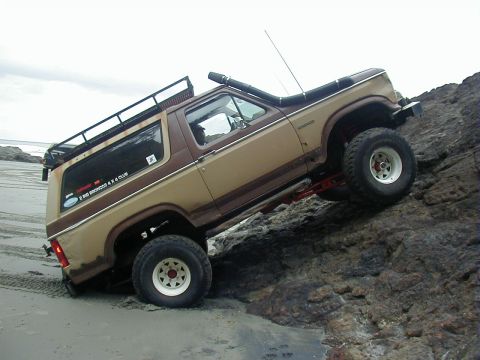 1994 Ford Explorer - This modified Ford Bronco navigates a rocky incline near the beach, equipped with off-road enhancements like a snorkel and roof rack.