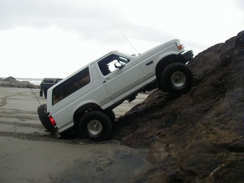 1994 Ford Explorer - White Ford Bronco navigating rocky terrain on a beach, with another vehicle in the distance.