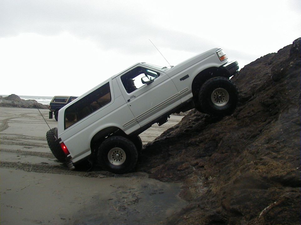 White Ford Bronco with lifted suspension and large tires climbing over rocks on a sandy beach, another dark SUV is visible in the background