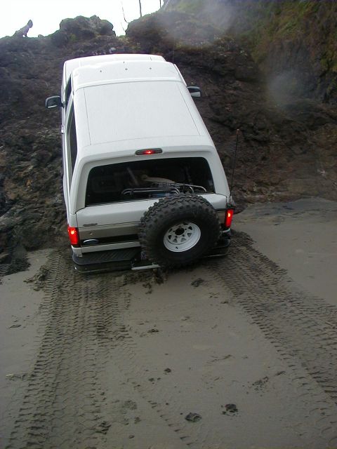 1994 Ford Explorer - A white SUV is stuck on a sandy beach next to a rocky cliff, with tire tracks indicating its struggle.