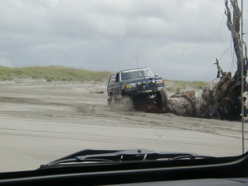 Dark blue SUV with off-road modifications driving on a sandy beach near a large driftwood log