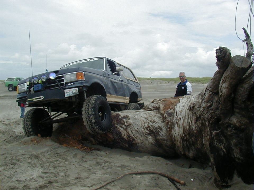 Dark blue lifted Ford Bronco with off-road tires on a sandy beach, climbing over a large driftwood log