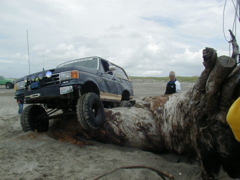 1994 Ford Explorer - A lifted dark blue SUV with off-road tires is shown navigating a large log on a sandy beach, with another vehicle visible in the background.