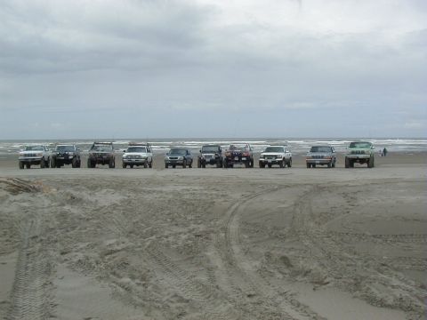 1994 Ford Explorer - A group of off-road vehicles are lined up on the sand near the ocean under a cloudy sky.
