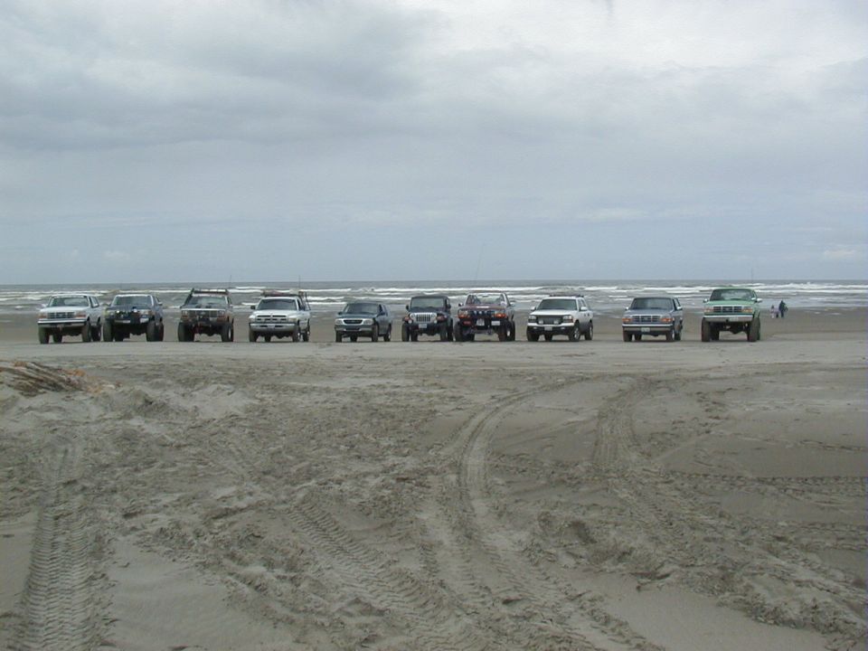 Row of ten lifted 4x4 trucks and SUVs parked on a gray overcast beach with surf behind