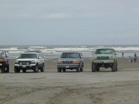 1994 Ford Explorer - Several trucks and SUVs are lined up on a beach with the ocean waves in the distance.