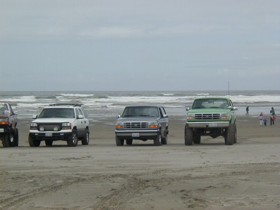 Three lifted 4x4 trucks lined up facing the camera on an ocean beach