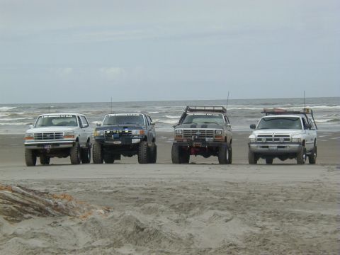1994 Ford Explorer - Four SUVs are parked on the sand near the ocean waves under an overcast sky.