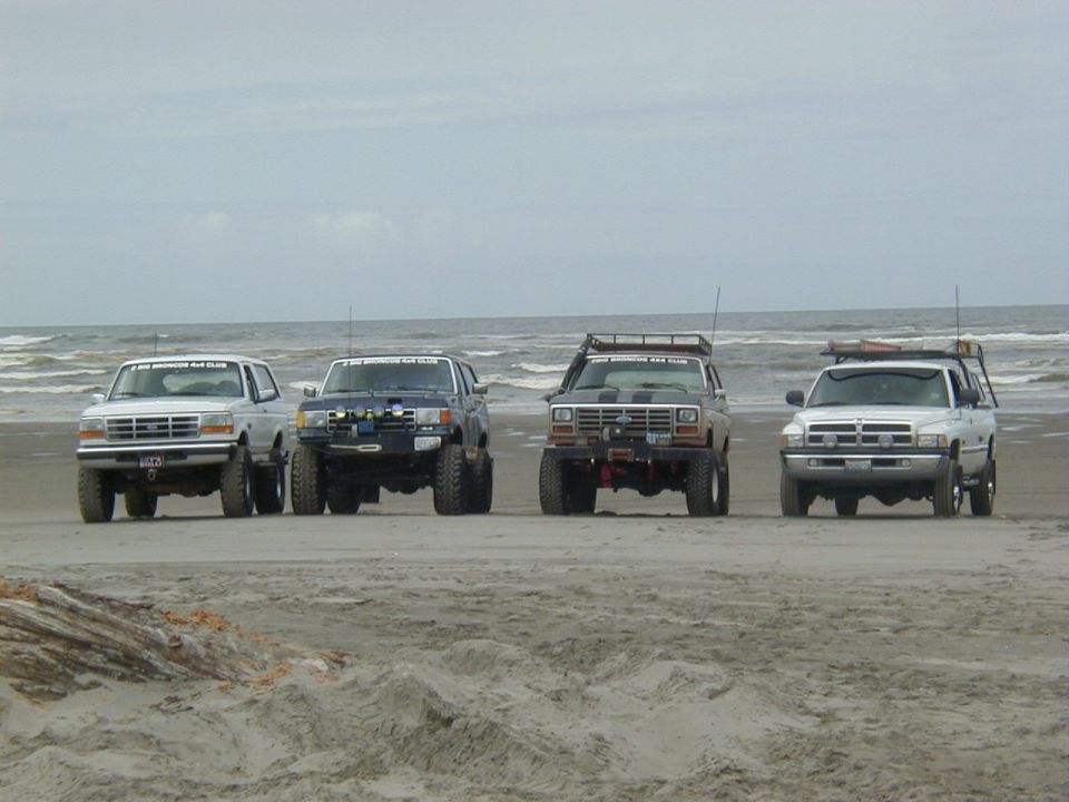 Four lifted 4x4 trucks and Broncos lined up on a sand beach with surf behind