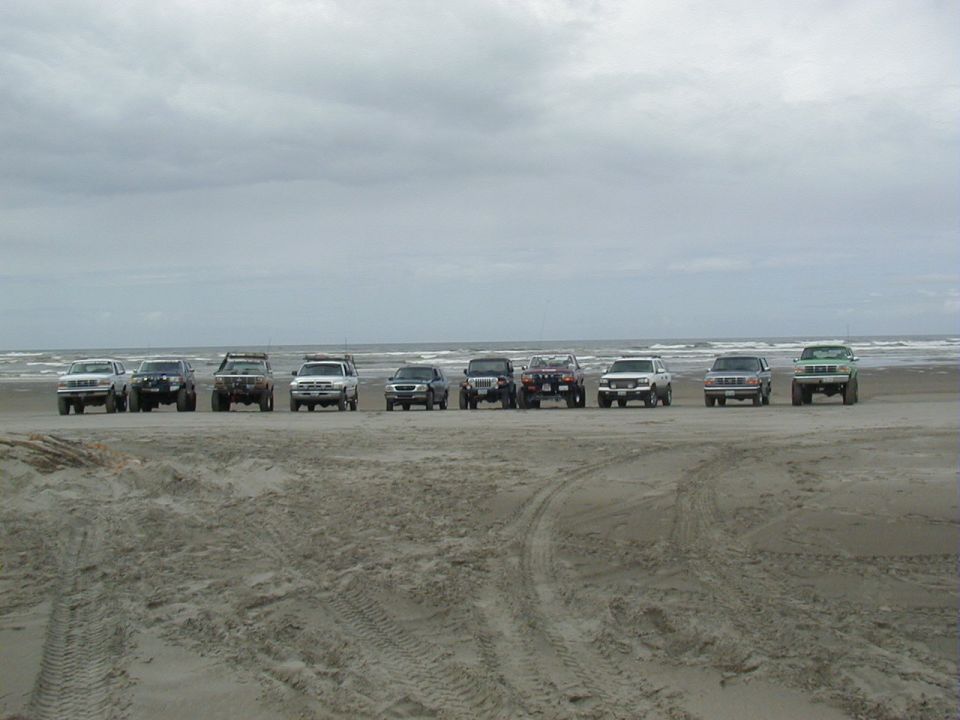 Ten 4x4 trucks and SUVs lined up facing the camera on an overcast beach