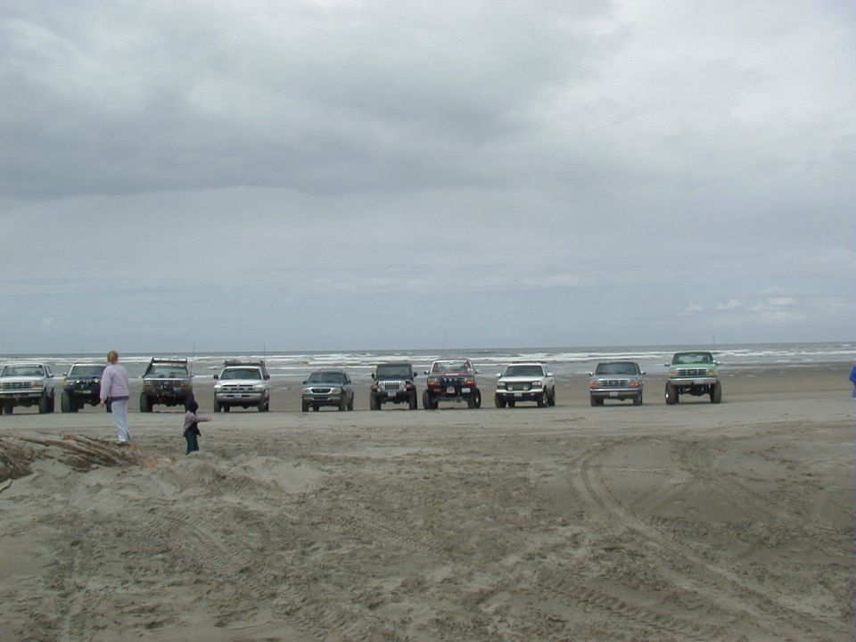 Line of SUVs and trucks on a beach under a cloudy sky