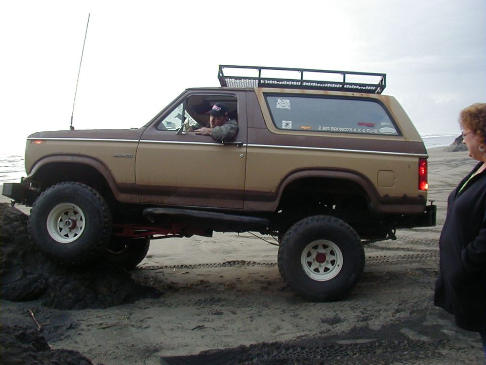 Brown and tan Ford Bronco with roof rack on sand, driver visible. A woman stands to the right