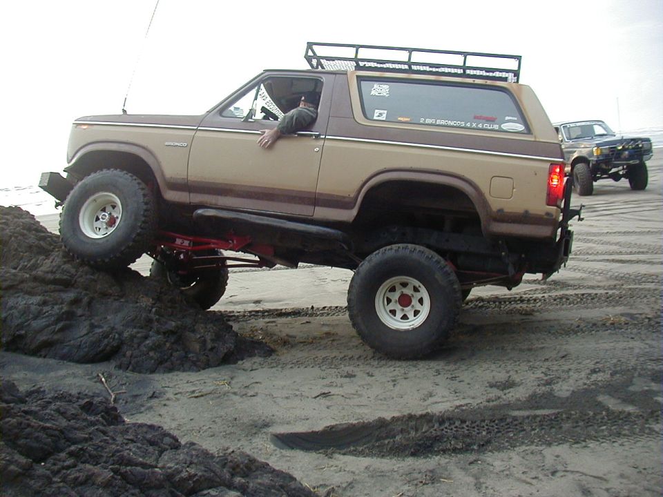 Brown and tan lifted Ford Bronco flexing over a peat bank on the beach with red radius arms