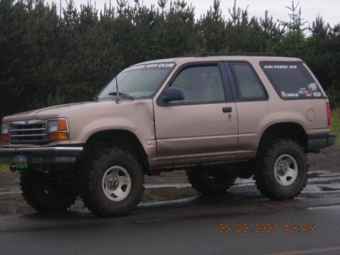 1994 Ford Explorer - A lifted 1994 Ford Explorer with off-road tires is parked on a wet surface, with trees in the background.