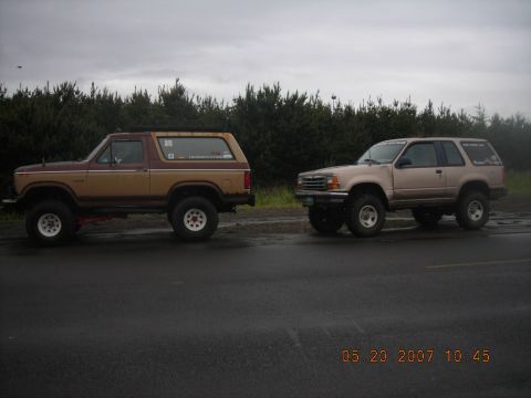 1994 Ford Explorer - Two lifted SUVs, a 1994 Ford Explorer and an older Ford Bronco, sit parked on a wet road.