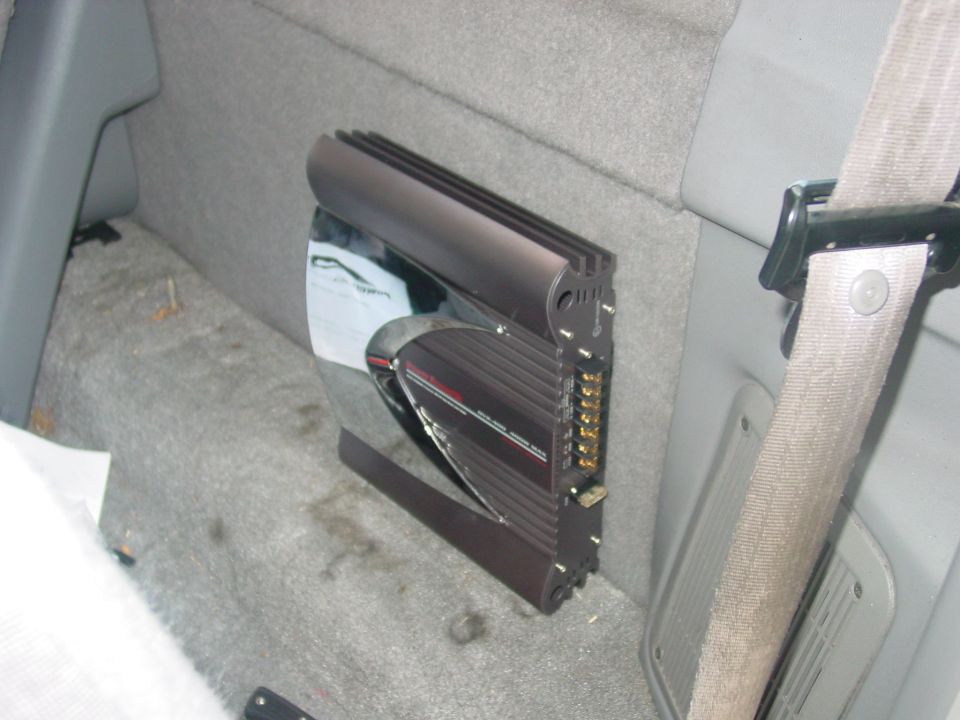 Car amplifier mounted on gray carpeted interior wall next to a seatbelt
