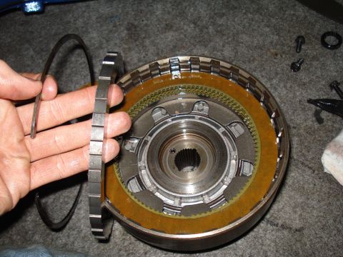 1984 Ford Bronco - A person is holding a metal ring above a disassembled transmission clutch pack, with tools and parts scattered around.