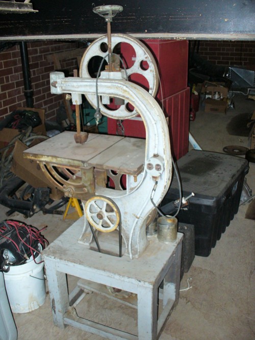 White bandsaw with a large wheel and a table, on a stand, in a cluttered room