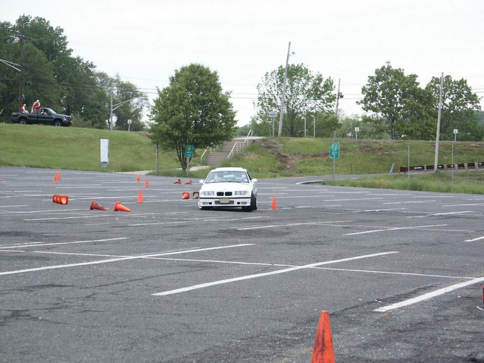 White BMW E36 coupe driving through cones in a parking lot. Black pickup truck with people in the background