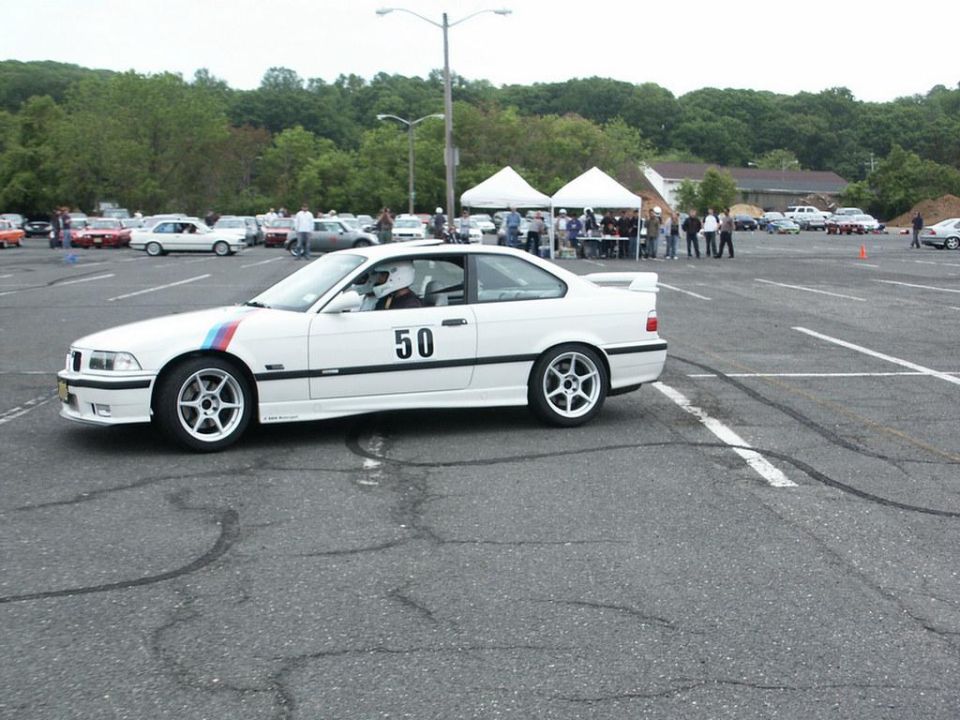 White BMW M3 coupe with racing stripes on fender and number 50 on door, parked in a lot with other cars and tents