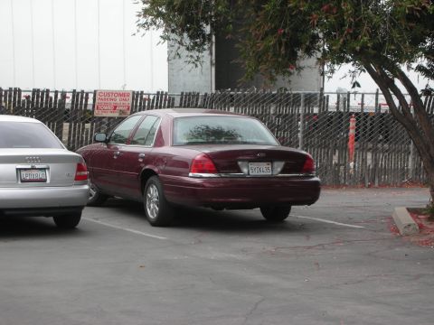 1992 Ford Crown Victoria - A maroon 1992 Ford Crown Victoria is parked next to a silver sedan, with a fence and trees in the background.