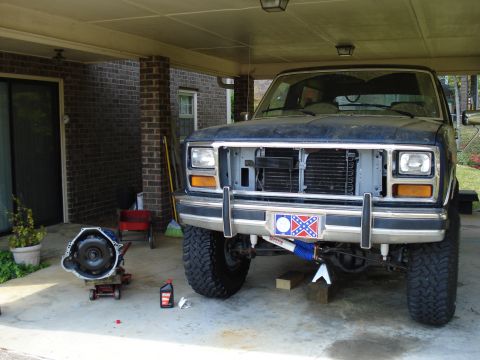 1984 Ford Bronco - A Ford Bronco is parked under a carport with its front grille removed and a transmission jack nearby, suggesting maintenance work.