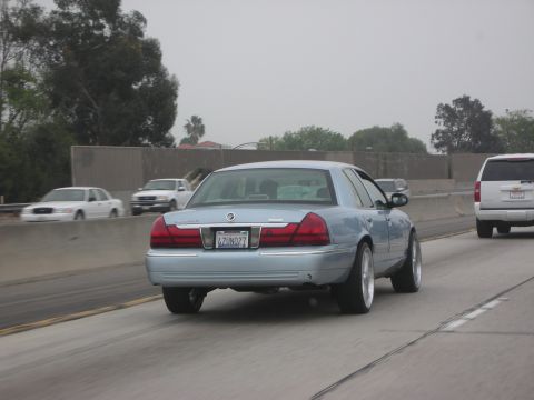 1998 Ford Crown Victoria - A light blue sedan drives on the highway, showing its rear and custom wheels.