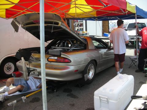 1998 Ford Crown Victoria - A silver sports car with racing modifications, including a roll cage, is parked under a canopy with its trunk open while a mechanic works u…