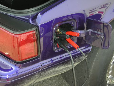 1998 Ford Crown Victoria - Jumper cables are connected to the fuel filler port of a purple vehicle, suggesting an unusual charging or power source.