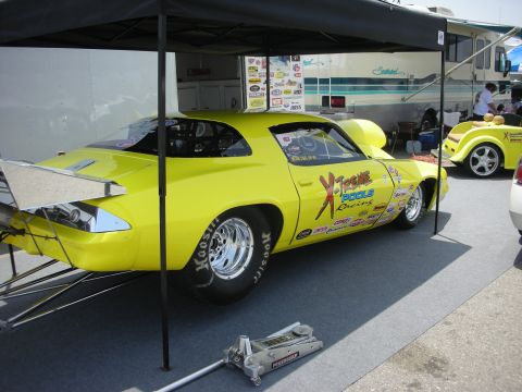 1998 Ford Crown Victoria - A bright yellow drag car, covered in various racing sponsorships, sits under a black canopy at what appears to be a race event.