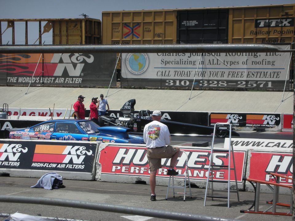 Two drag cars are lined up at a drag strip. A man in a white shirt watches from a stool