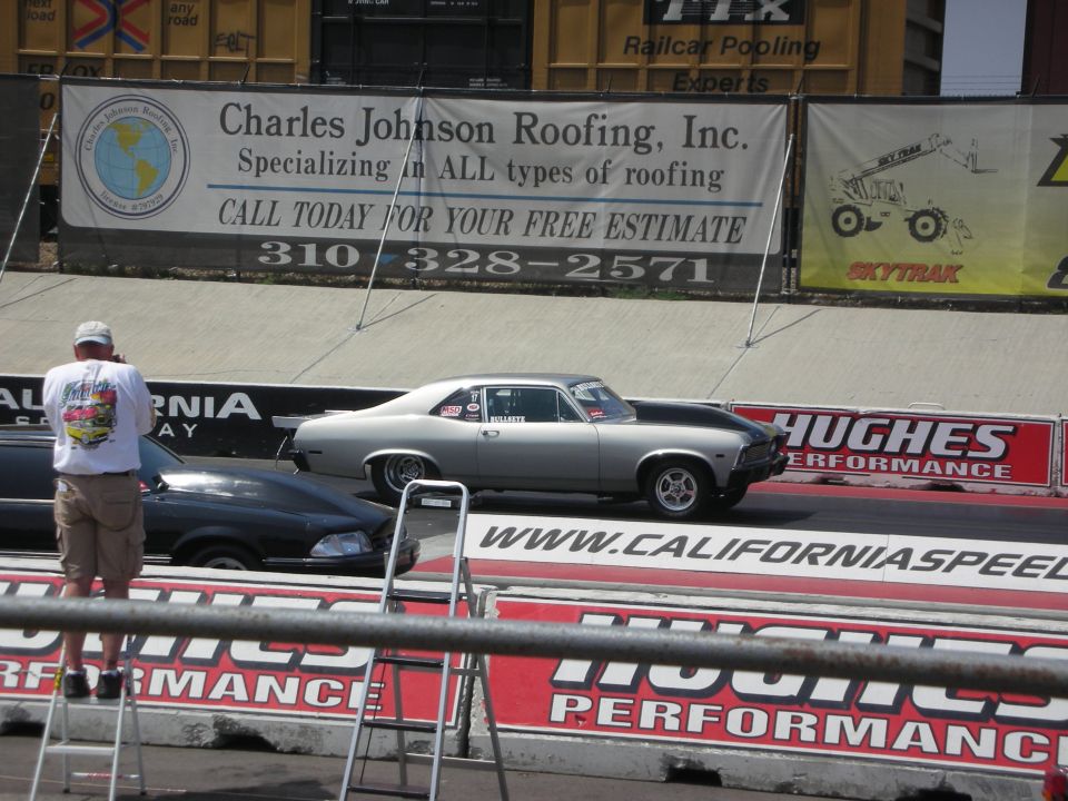 Silver classic coupe with a black hood on a race track, another car is to its left. A man stands on a ladder taking a photo