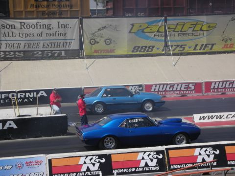 1998 Ford Crown Victoria - Two cars compete on a drag strip with advertising banners in the background.