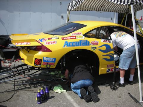 1998 Ford Crown Victoria - Mechanics work on the underside of a yellow drag racing car during a pit stop.