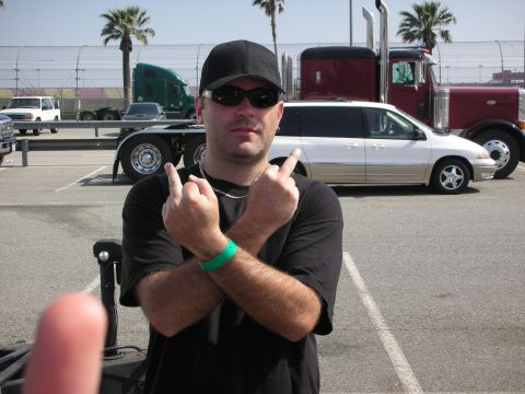 1998 Ford Crown Victoria - A man stands with his arms crossed, giving the finger, with large trucks and a minivan parked behind him.