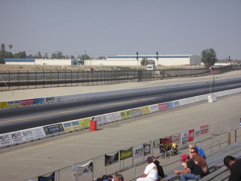 1998 Ford Crown Victoria - A drag racing track is shown with spectators in the stands and buildings in the distance under a clear sky.