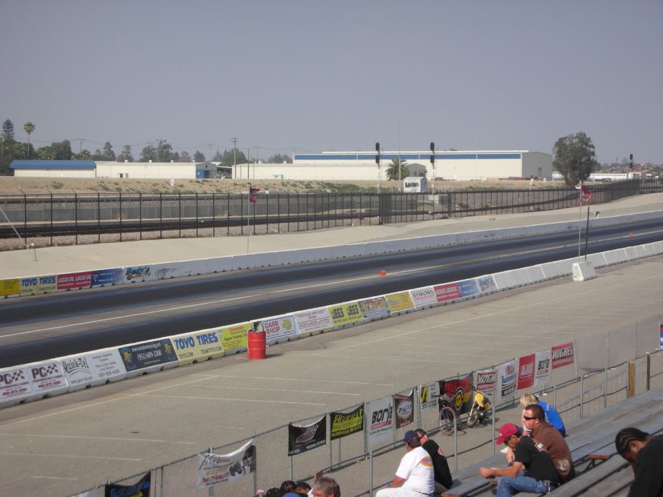 View of a drag strip with spectator seating in the foreground and industrial buildings in the background