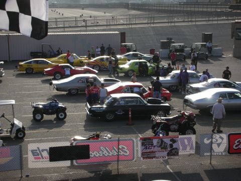 1998 Ford Crown Victoria - A variety of vehicles are gathered at a race track, with people milling around.