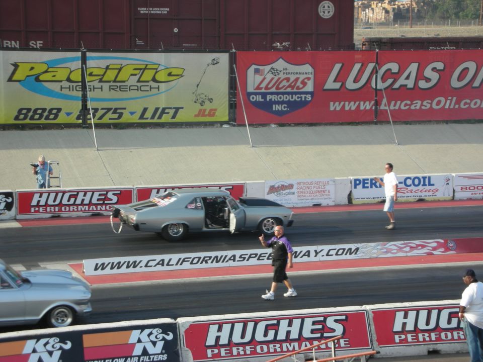 Drag strip with a silver muscle car and another car, spectators, and sponsor banners