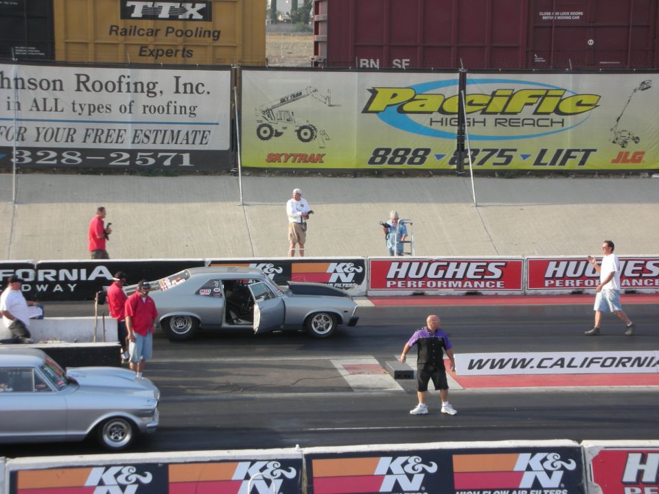 Two cars on a drag strip, one silver with doors open. People around