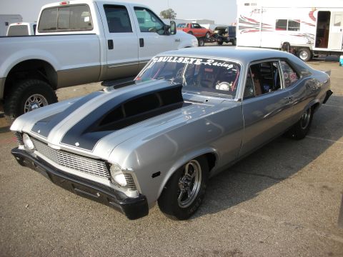 1998 Ford Crown Victoria - Silver 1969 Chevrolet Nova drag car features a prominent hood scoop and racing decals, seen with other vehicles in the background.