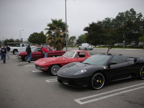 1998 Ford Crown Victoria - A black convertible sports car is parked in a lot alongside several other vehicles, including a red classic car and a white pickup truck.