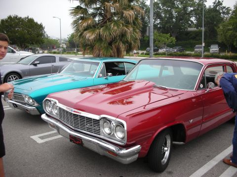1998 Ford Crown Victoria - A red classic car with a prominent hood scoop is parked next to a teal classic car.